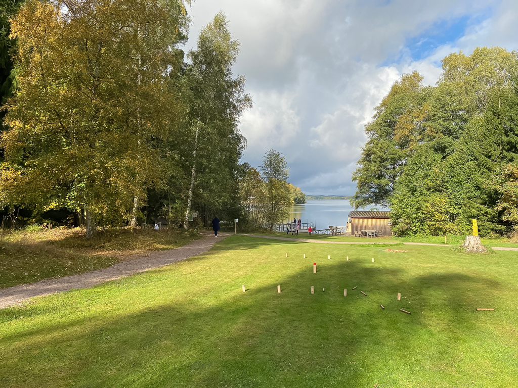 Gräsplan med kubbspel uppställt, omgiven av träd och med en sjö i bakgrunden.